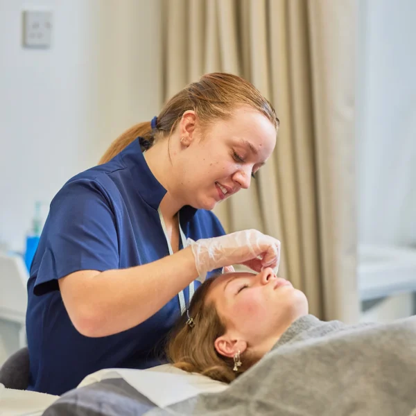 A woman lies on a treatment bed while a technician examines her face.