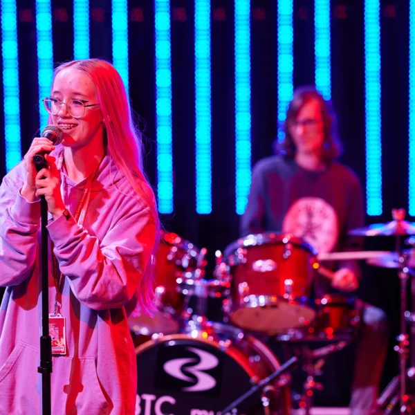 A female singer performing on stage with a male drummer in the background. The singer is holding a microphone and wearing a pink jacket.