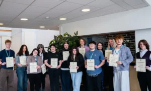 A group of young people proudly holding certificates and wearing blue lanyards, standing in a well-lit room with a plant in the background.