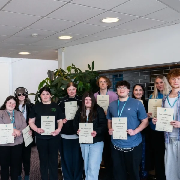 A group of young people proudly holding certificates and wearing blue lanyards, standing in a well-lit room with a plant in the background.