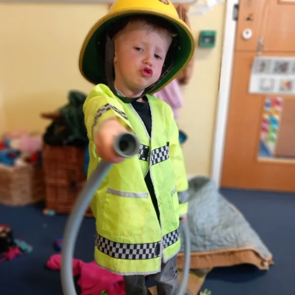 Young child wearing a fireman's hat and vest holding a toy hose.