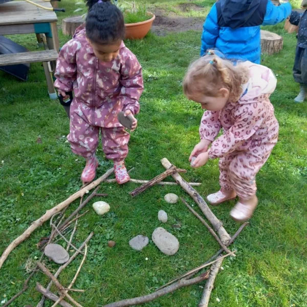 Two young girls in pink tracksuits building a circle with sticks and stones on grass.