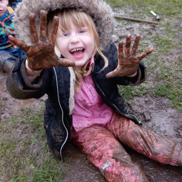 A young girl with blonde hair covered in mud sitting on the ground with her hands up.