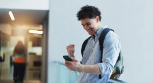 A young man with a backpack on looks at his phone with a clenched fist in excitement.