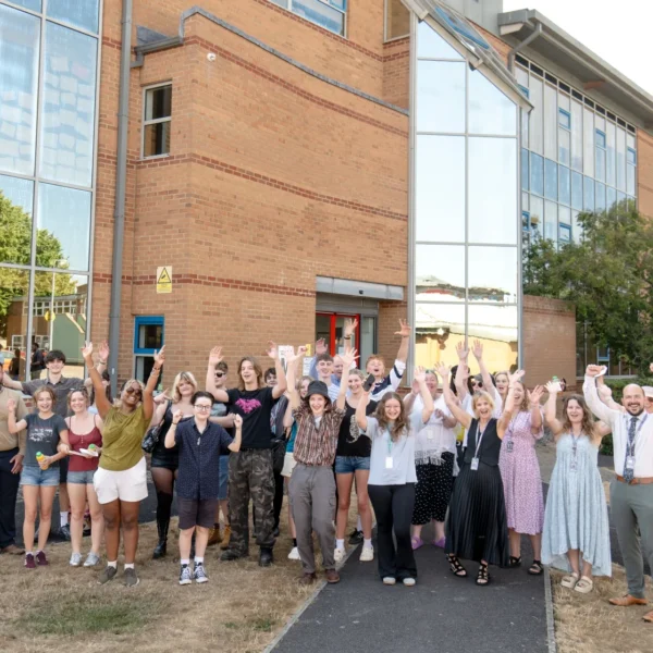 GROUP OF STUDENTS CHEERING OUTSIDE A BRICK BUILDING WITH GLASS WINDOWS.