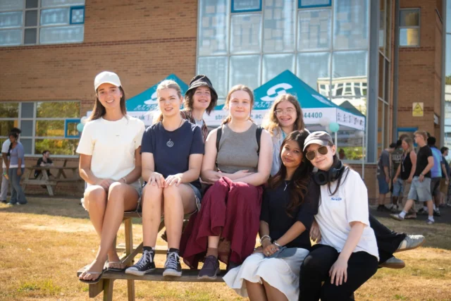 A group of young people sitting on a bench outside a modern building, smiling for the camera.