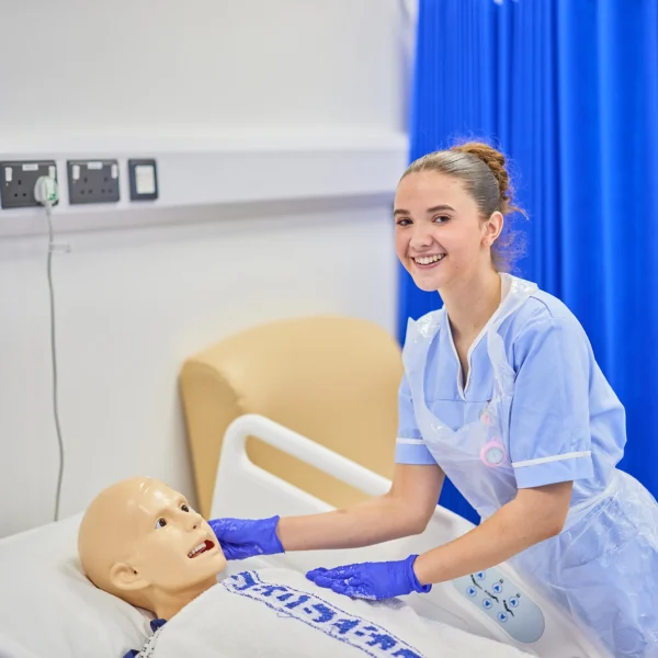 A woman in blue scrubs interacts with a mannequin on a hospital bed.