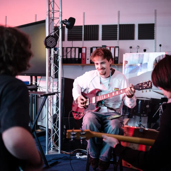 Man playing electric guitar in a dimly lit room with two people watching.