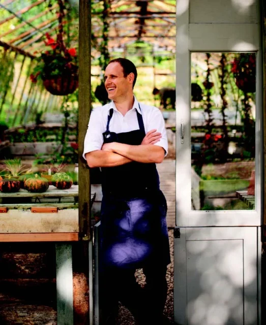 Man in a black apron standing in the doorway of a greenhouse.
