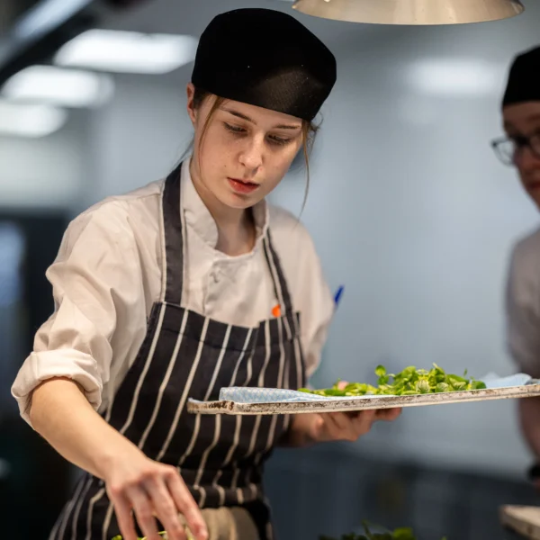 Chef garnishing a dish with greens in a kitchen.