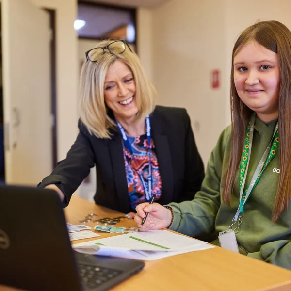 A teacher and student sitting at a desk with a laptop and papers.