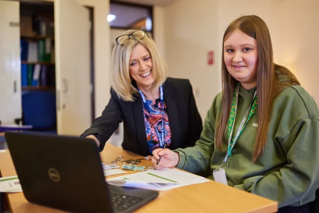 A teacher and student sitting at a desk with a laptop and papers.