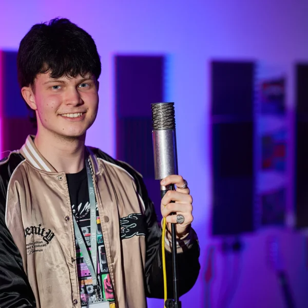A young man holding a microphone in a recording studio with purple and blue lighting.