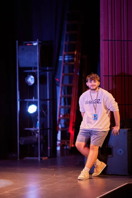 A man on a stage with a lanyard around his neck, wearing a white t-shirt and shorts.