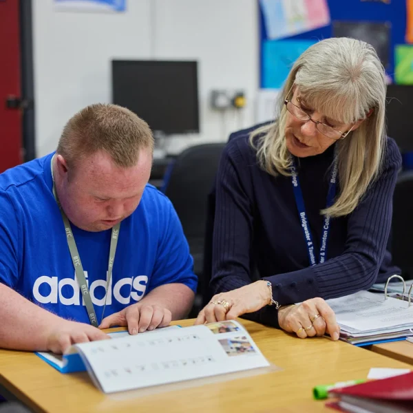 A woman supporting a man with a disability at a desk.