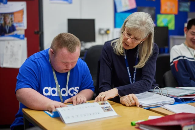 A woman supporting a man with a disability at a desk.