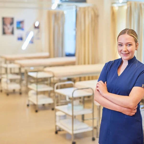 A smiling woman in blue nurse scrubs standing in a hospital room with medical equipment