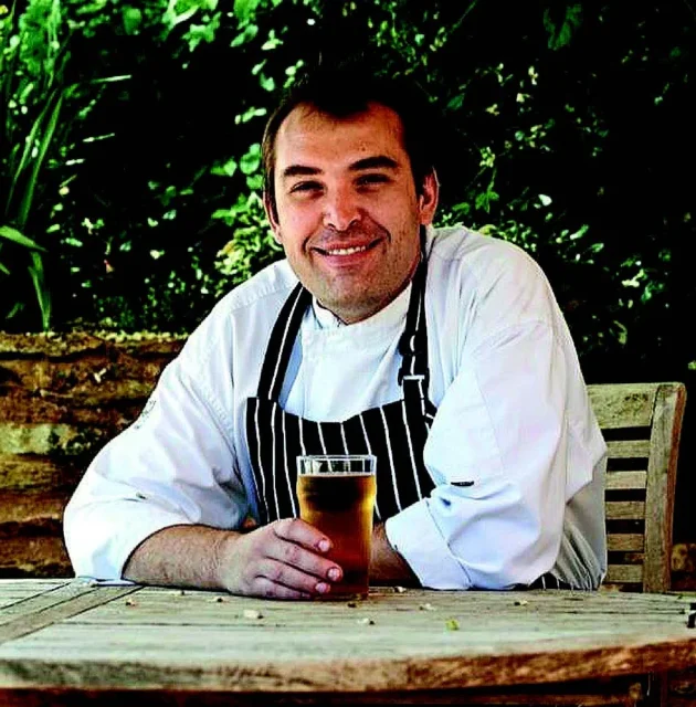 Smiling chef in a white shirt and black apron holding a glass of beer on a wooden table outdoors.