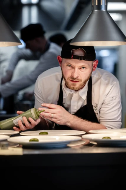Chef preparing food in a kitchen, garnishing a dish with a green sauce.