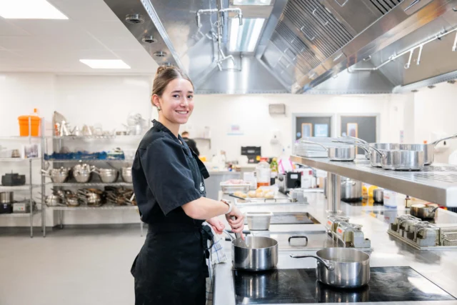 A woman in a kitchen with pots and pans on the stovetop. She appears to be a chef or cook.