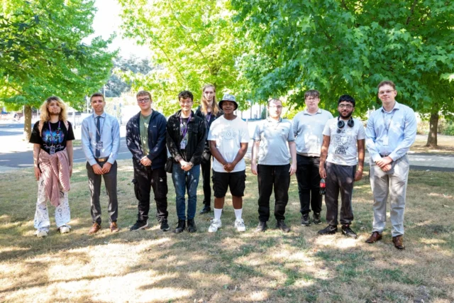 A group of young people standing outdoors in a park-like setting, with green trees and grass providing a natural backdrop.