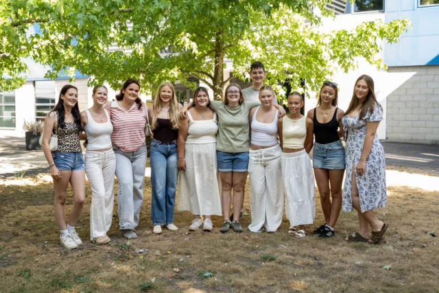 A group of young people smiling and posing outside under a tree, dressed in casual summer attire.