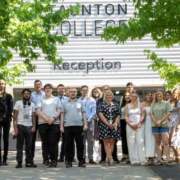 A group of students and staff posing in front of Taunton College's reception building, framed by green foliage.