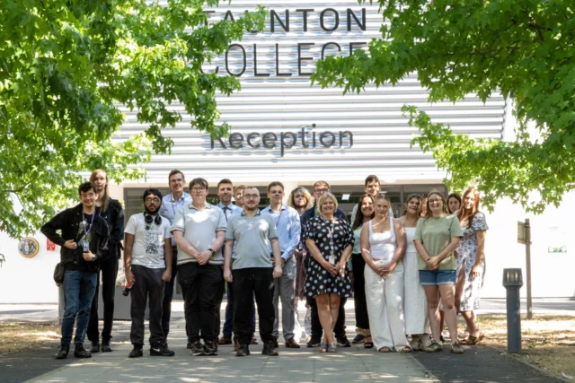 A group of students and staff posing in front of Taunton College's reception building, framed by green foliage.
