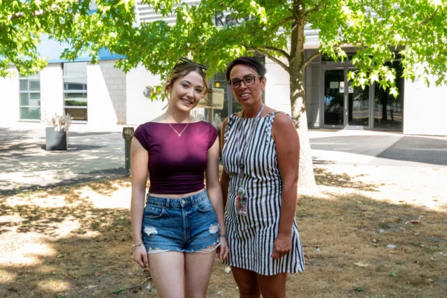Two women standing outdoors under a tree, one in a purple top and denim shorts, the other in a striped dress and glasses.