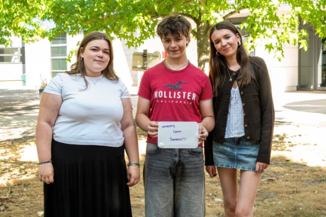 Three young people standing outdoors holding a sign celebrating academic success or school event